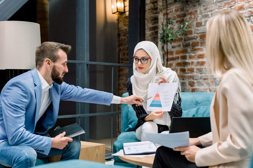 Multiethnic business people talking during meeting in office. Young Caucasian man explaining proposal. Pretty Muslim businesswoman showing marketing strategy on paper to colleague